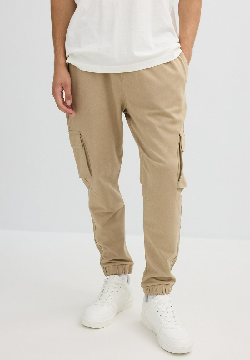 Hombre vistiendo pantalones jogger beige tipo cargo, zapatillas blancas y una camiseta blanca, de pie con las manos en los bolsillos frente a un fondo liso.