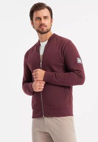 Bordeaux zippered bomberjakke med ribbede ærmer og kant, med et brystpatchlogo og sidelommer, lavet af blødt stof.