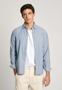 Lichtblauwe overhemden met een zachte textuur, lange mouwen opgerold bij de manchetten, gedragen over een wit t-shirt, gecombineerd met lichtgekleurde broek.