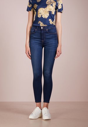 Kvinde iført mørkeblå skinny jeans, marineblå floral kortærmet top og hvide sneakers, stående mod en ensartet lys baggrund.