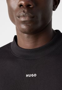 Felpa nera con colletto a costine e logo bianco "HUGO" sul davanti. Il tessuto ha una consistenza liscia e una vestibilità rilassata.