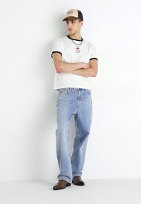 White t-shirt with black trim, red logo at chest; light blue jeans; brown shoes; beige cap with brand logo; simple, relaxed fit.