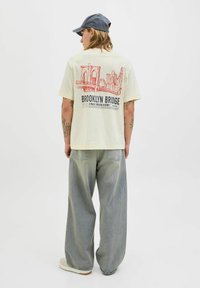 Lys beige t-shirt med korte ærmer, der har et rødt Brooklyn Bridge grafik på ryggen, sammen med løse lysegrå jeans og beige sneakers.