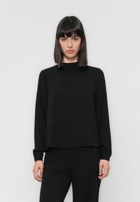 Black long-sleeve top with a high, gathered neckline and textured fabric. Relaxed fit with slightly loose sleeves, worn with black trousers.