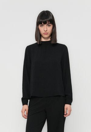 Black long-sleeve top with a high, gathered neckline and textured fabric. Relaxed fit with slightly loose sleeves, worn with black trousers.