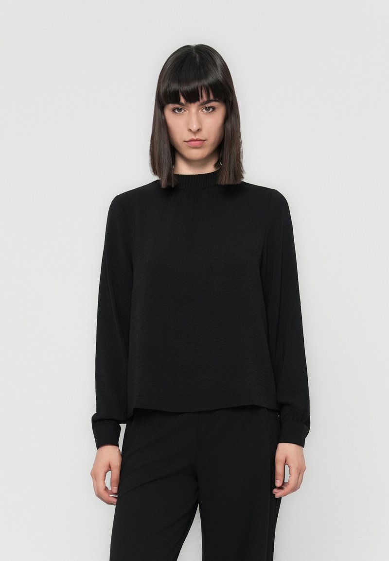 Black long-sleeve top with a high, gathered neckline and textured fabric. Relaxed fit with slightly loose sleeves, worn with black trousers.