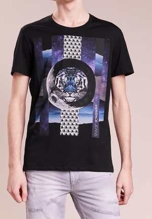 T-shirt noir en coton avec un motif représentant un tigre, une lune et des motifs géométriques dans diverses couleurs, avec "VERSACE JEANS" imprimé sur le côté.