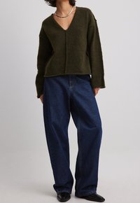 Olijfgroene gebreide sweater met een diepe V-hals, gecombineerd met losse blauwe denim jeans. De sweater heeft een recht model en zichtbare naden.