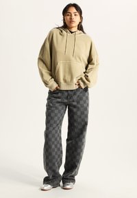 Beżowy, oversize'owy hoodie z przednią kieszenią i sznurkiem, w zestawieniu z szaro-czarnymi, w kratę szerokimi spodniami i srebrnymi butami.