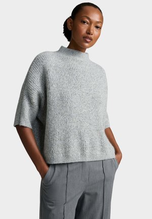 Donna con i capelli tirati indietro che indossa un maglione smanicato leggero grigio chiaro con texture e pantaloni grigi plissettati con le mani nelle tasche.