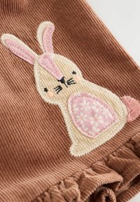 Teddystof med påsyet beige og lyserød kaninapplikation, der har broderet ansigt, blomsterprydet mave og en fluffy lyserød hale.