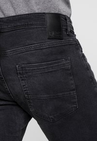 Black denim jeans feature a fitted design with a visible back pocket and a black leather brand label at the waistband.