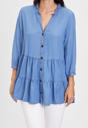 Kobieta ubrana w jasnoniebieską, warstwową bluzkę na guziki z rękawami 3/4 i falbankowanym kołnierzem, w komplecie z białymi spodniami.