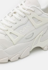 Fehér sneaker hálós és bőr anyagokból. Vastag talppal, textúrázott díszítésekkel és kivágott részletekkel rendelkezik, letisztult, modern dizájnban.