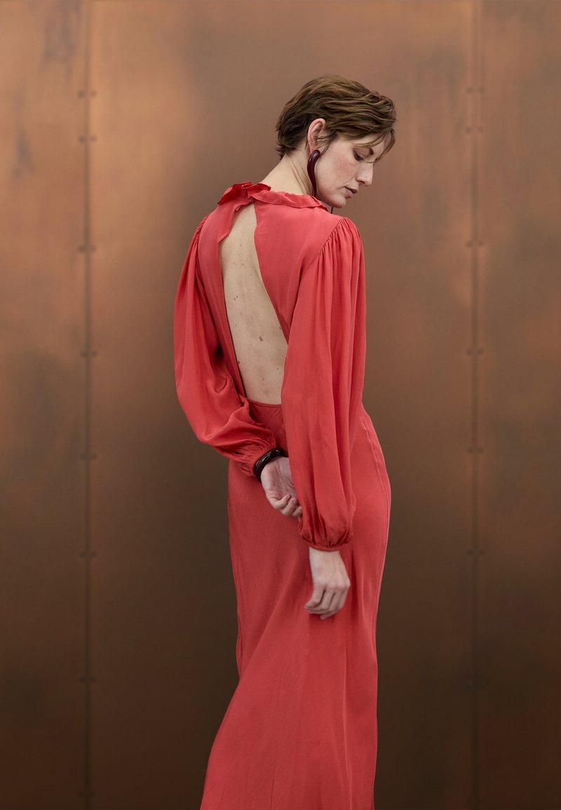 Ilga raudona suknelė su aukštu kaklu, atvira nugara ir pūstomis ilgomis rankovėmis. Pagaminta iš švelnios medžiagos, su raukiniais riešų srityje.