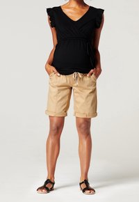 Haut enveloppant noir avec des manches courtes à volants, associé à un short beige retroussé avec cordon de serrage. Des sandales plates noires complètent le look.