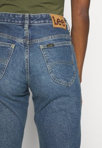 Blauwe denim jeans met een hoge taille, voorzien van een bruine leren Lee merkpatch, gele stiksels en standaard achterzakken.