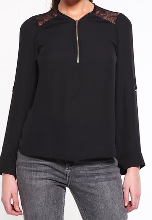 Mujer con una blusa negra de manga larga con detalles de encaje en los hombros y cremallera frontal, combinada con vaqueros grises.