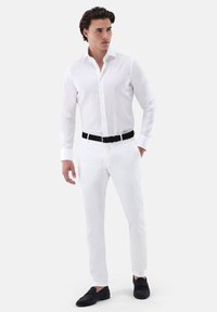 Weißes Langarmhemd mit einem Button-Down-Kragen, kombiniert mit weißen Slim-Fit-Hosen und schwarzen Mokassins, dazu ein schwarzer Ledergürtel.