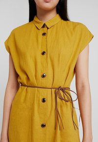 Šaty ve hořčicově žluté barvě vyrobené z lehké tkaniny, s knoflíkovým zapínáním vpředu, krátkými rukávy a hnědým pleteným páskem v pase.