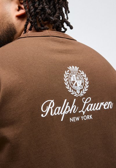 Κοντινή λήψη ατόμου που φορά καφέ πουκάμισο με λευκό λογότυπο Ralph Lauren New York και έμβλημα στην πλάτη, με κοντά στριφτά μαλλιά εμφανή.