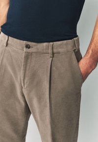 Chinos marron clair avec une texture lisse, des plis sur l'avant et une fermeture par bouton. Associés à un haut ajusté bleu marine.