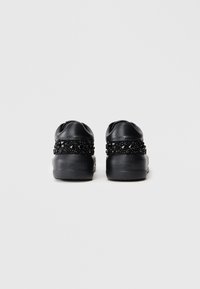Sapatilhas pretas com parte traseira em tecido texturizado e adornadas com gemas pretas. Design de bico arredondado, apresentando uma sola de borracha plana com marca.