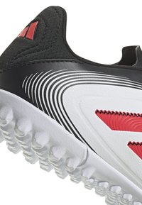 Zapatilla deportiva en blanco y negro con acentos rojos, parte superior texturizada, lateral de cuero liso y suela de goma translúcida y con patrón para mayor tracción.