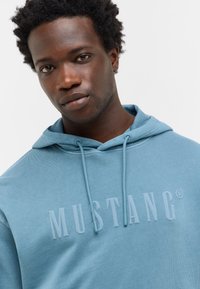 Gros plan sur un homme aux cheveux bouclés courts portant un sweat à capuche bleu clair avec "MUSTANG" imprimé sur le devant.