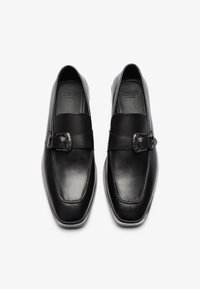 Mocasinés de cuero negro con una textura suave, que cuentan con una correa frontal y un detalle de hebilla plateada. Diseño de punta cuadrada y perfil minimalista.