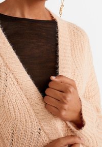 Ylipitkä, neulottu neuletakki pehmeän vaalean persikan värisenä, jossa on syvä V-pääntie ja pörröinen pinta; kerroksittain tiukan mustan paidan päällä.