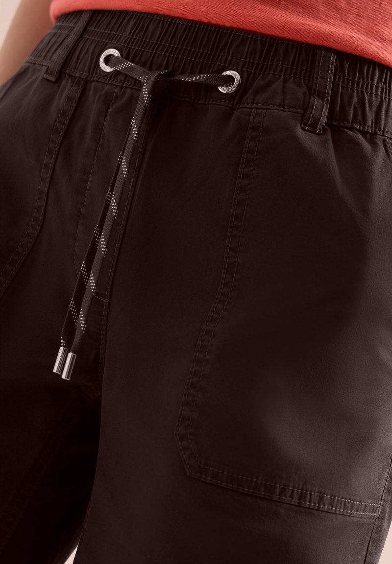 Short noir à taille élastique avec cordon de serrage et poche avant visible, porté avec une chemise rouge rentrée légèrement au-dessus de la taille.