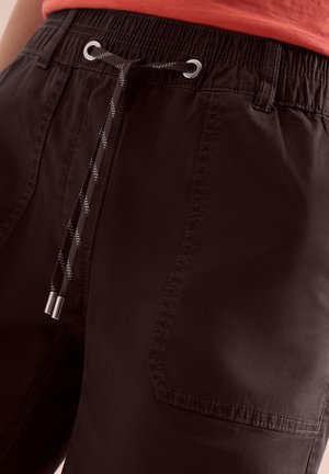 Short noir à taille élastique avec cordon de serrage et poche avant visible, porté avec une chemise rouge rentrée légèrement au-dessus de la taille.