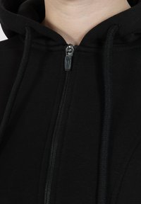 Sudadera con cremallera negra de tejido texturizado, con un cuello alto y cordones, resaltada por una cremallera metálica y detalles en negro.