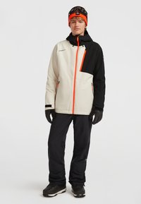 Un jeune homme portant une veste de ski bi-colore beige et noire avec des fermetures éclair orange, un pantalon de ski noir, des gants, un bonnet orange et des lunettes de ski sur la tête.