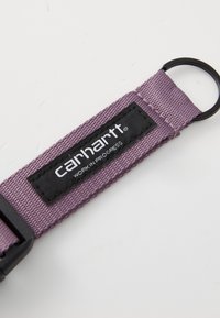 Lilla nylonrem med en tekstureret overflade, der har et sort label med teksten "carhartt WORK IN PROGRESS" og en cirkulær metalkrog.