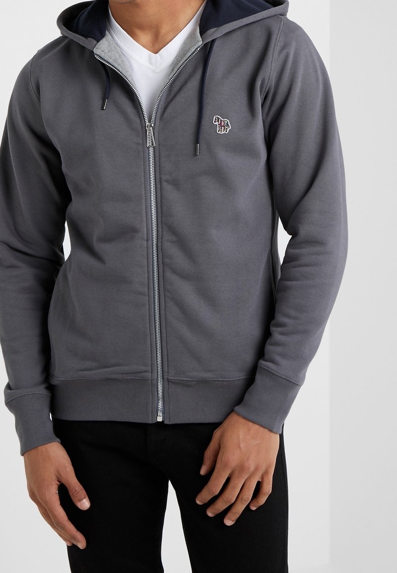 Homme portant un sweat à capuche gris zippé avec un petit patch brodé sur la poitrine, par-dessus un t-shirt blanc à col en V et un pantalon noir.