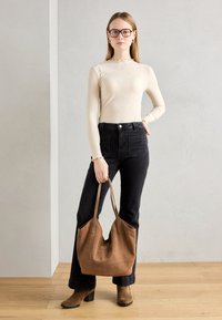 Bolso tote de ante marrón con asas planas, combinado con una camiseta de canalé crema y jeans negros de cintura alta, sobre un fondo gris claro.