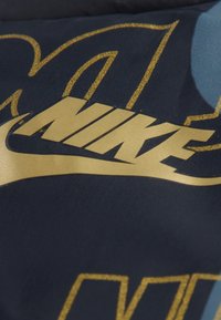 Nike logo u zlatnoj boji, na teksturiranoj tkanini s mornarskom pozadinom. Podebljana slova sadrže zakrivljene dizajnerske elemente i akcent povijanja.