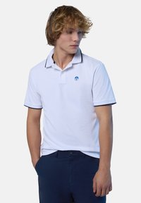 Polo bianca con bordature scure, maniche corte, due bottoni al colletto e un logo circolare sul petto sinistro. Il tessuto sembra liscio.