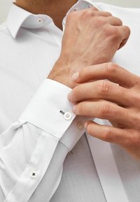 Biała koszula z guzikiem o teksturowanej tkaninie, zbliżenie na mankiet z guzikiem i małym zielonym akcentem nici.