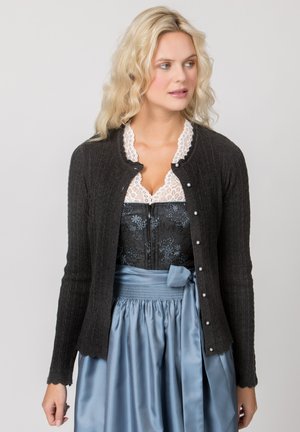 Blonde Frau trägt einen schwarzen, geknöpften Strickjacke über einer weißen Spitzenbluse und einem schwarzen floralen Korsett mit einem blauen Satinsrock, der an der Taille gebunden ist.