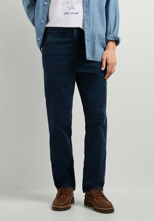 Mann trägt dunkelblaue Hose, braune Lederschuhe, weißes Grafik-T-Shirt und blau gestreiftes Hemd mit hochgekrempelten Ärmeln.