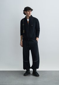 Μαύρο ριμπ ζακέτα με φερμούαρ και ψηλό γιακά, σε συνδυασμό με φαρδιά μαύρη παντελόνια με ανοιχτό γκρι ραφές και μαύρα αθλητικά παπούτσια.