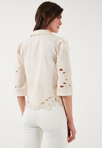 Camisa color crema con mangas abullonadas de tres cuartos, que presenta intrincados bordados de flores recortadas a lo largo de las mangas y el dobladillo.