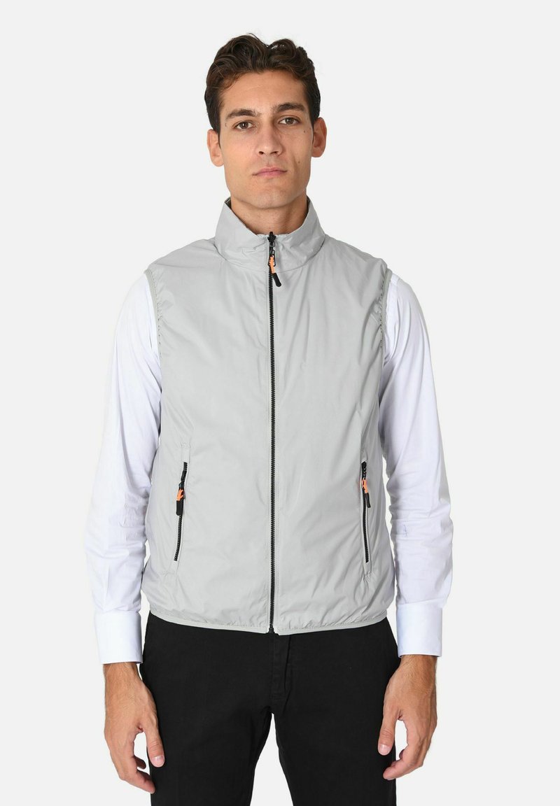 Uomo che indossa un gilet grigio chiaro senza maniche con zip sopra una camicia bianca a maniche lunghe e pantaloni neri, in piedi contro uno sfondo neutro.