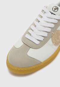 Hvit og lys grå sneaker med en semsket finish, som har hvite snørebånd, en gummisåle i brunfarge, og et rundt logo-patch på siden.