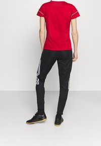 Červené sportovní tričko s bílými detaily, spárované s černými kalhotami s bílými pruhy po stranách. Černé sportovní boty s gumovou podrážkou.