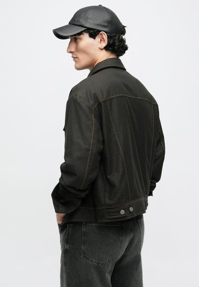 Jachetă din denim verde închis cu cusături portocalii contrastante, cu butoane tip snap și o croială lejeră, asortată cu pantaloni din denim negri și o șapcă.