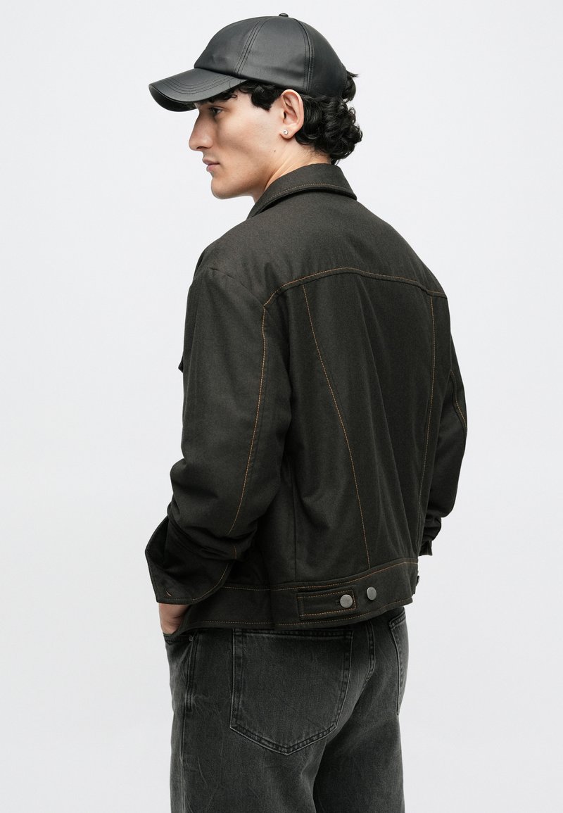 Mørkegrøn denimjakke med kontrasterende orange syninger, der har trykknapper og en afslappet pasform, parret med sorte denimbukser og en kasket.